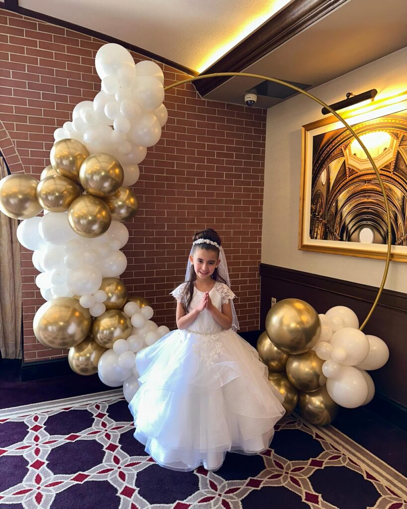 First Communion celebration featuring a white and gold balloon garland with a circular backdrop, designed by Caliella.