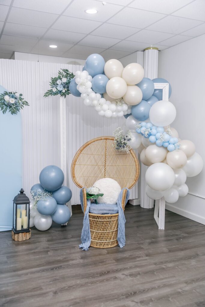 Elegant baby shower setup featuring pastel blue and white balloon garlands, rattan chair, and greenery by Caliella of RI.